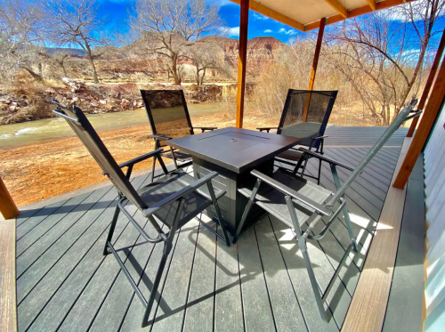 A patio with a black table and four chairs, overlooking a river and surrounded by bare trees and red rock formations.